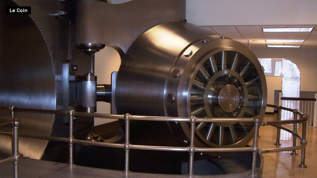 A massive round steel bank vault door stands open behind a polished metal guard rail, its spoked locking wheel facing the camera, in a brightly lit room.